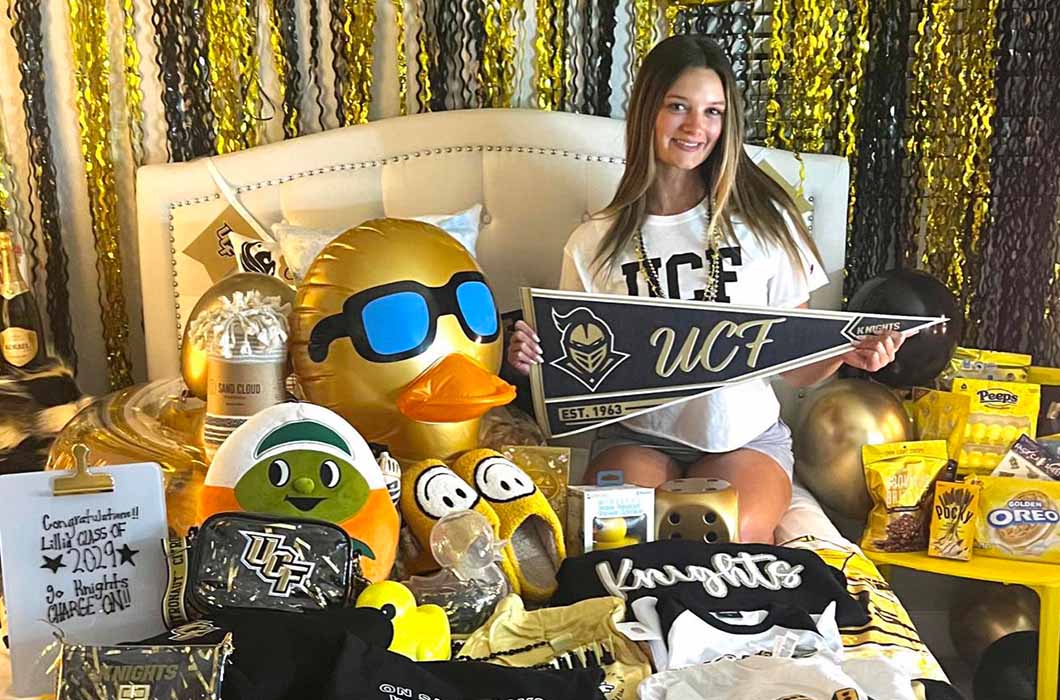 A young woman smiles on a bed covered in UCF Knights merchandise, including a pennant, plush toys, T-shirts, snacks, and decorations in black, gold, and white. A UCF banner and festive streamers are in the background. 