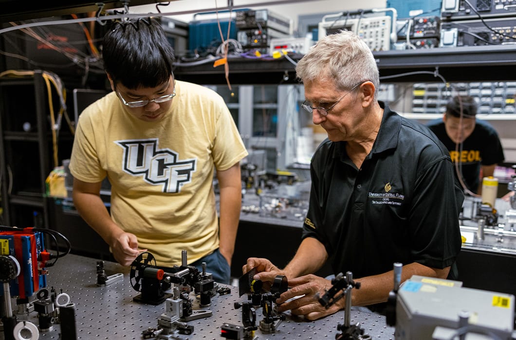 Two people work together on a lab bench filled with scientific equipment and optical devices. One wears a UCF t-shirt, and the other wears a black shirt with lab logos. Another person works in the background.