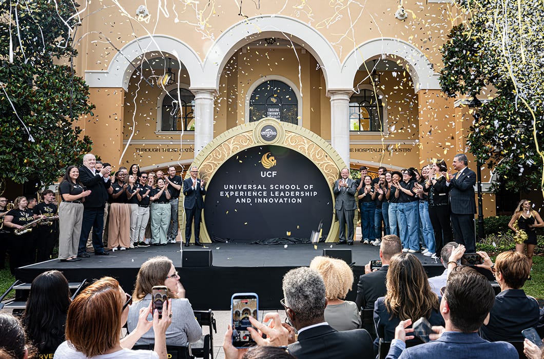 A large group of people stand on an outdoor stage under falling confetti at a UCF event, with an audience seated in front, and a backdrop reading Universal School of Experience Leadership and Innovation.