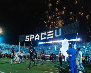 Football players run onto a brightly lit field under a large SPACE U banner, with fireworks lighting up the night sky and smoke effects adding excitement. The stadium is filled with cheering fans. 