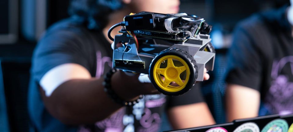 A UCF Knight Hacks member holds a small robotic vehicle with a yellow wheel, exposing its electronic components.