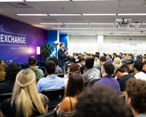 A speaker addresses a large, seated audience in a brightly lit room with a purple wall displaying the word EXCHANGE. People are attentively listening, and balloons are visible in the background.