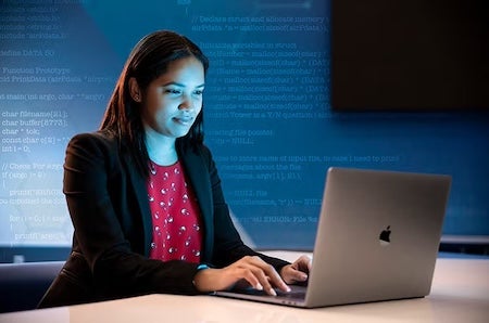 A woman in a blazer and red blouse types on a laptop at a desk, with blue computer code projected on the wall behind her, suggesting ai technology or programming environment.