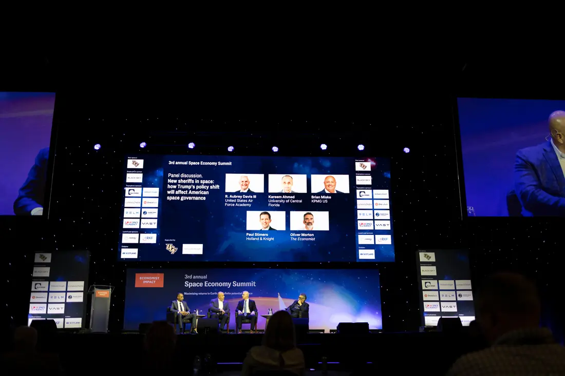 Panelists seated on stage in front of a large screen reading "Space Economy Summit," addressing a full arena audience at the Economist Space Economy Summit in 2025.