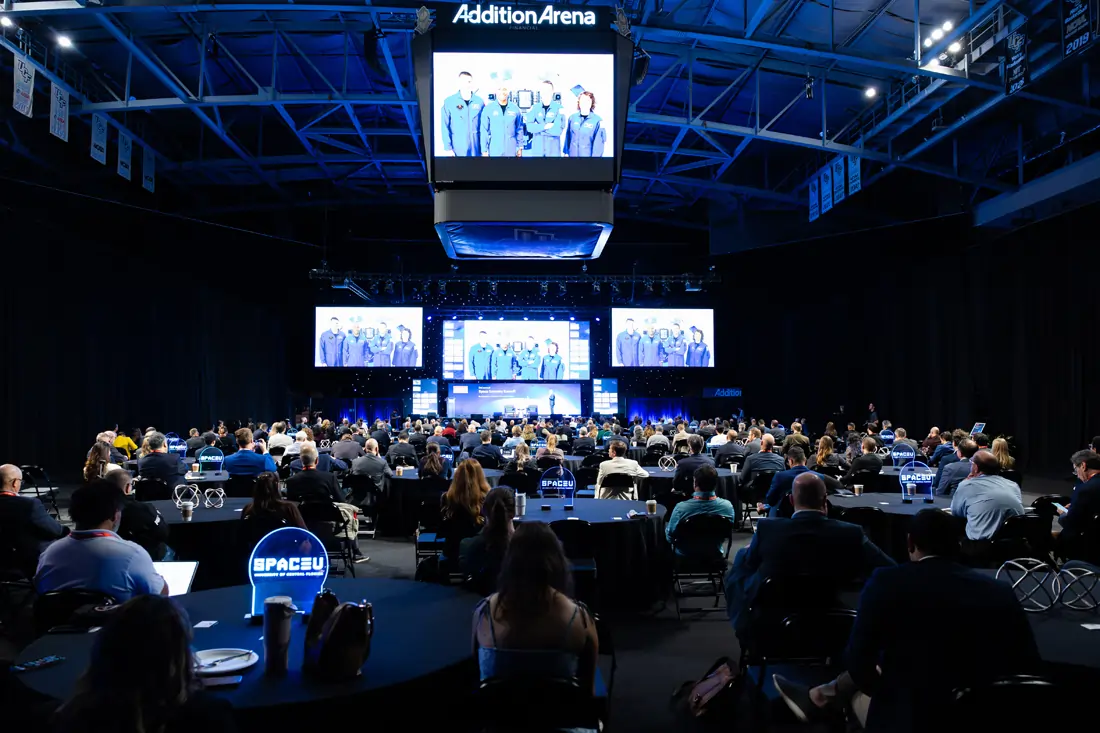 Attendees seated at round tables in a large arena watching presentations on overhead screens at the Economist Space Economy Summit in 2025.