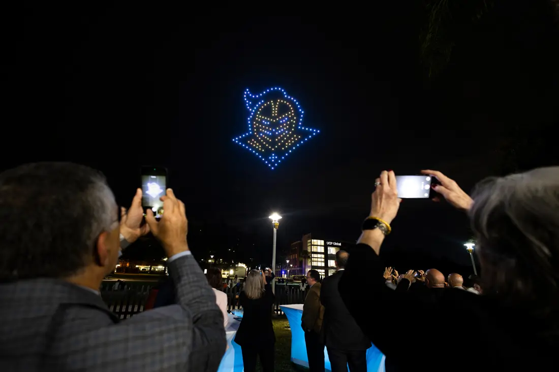 Guests holding up phones to photograph a glowing Knightro created by a drone light show.
