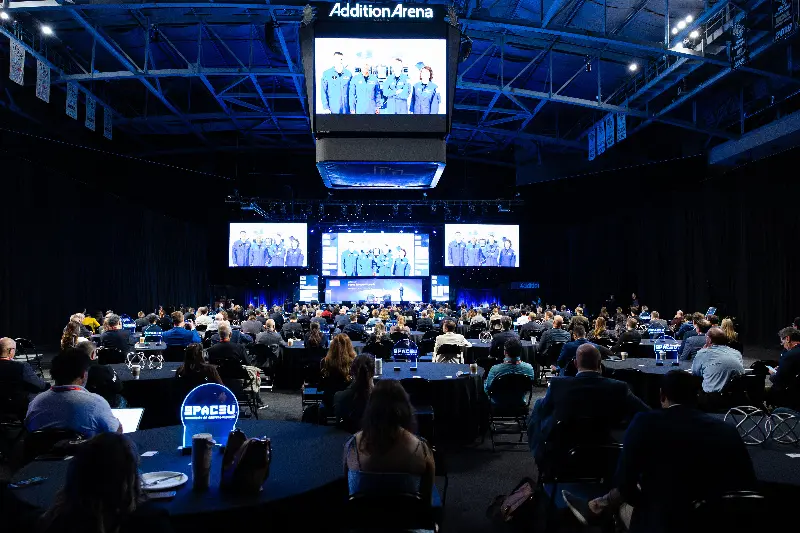 A speaker presents on stage to a large seated audience during the 2025 Space Economy Summit. Four large screens display an image of a crew of astronauts.