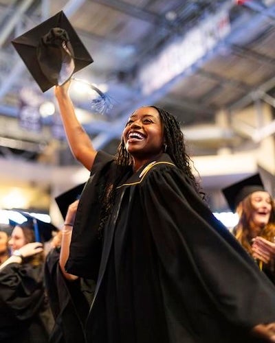 A joyful UCF graduate in a black cap and gown raises her cap high, smiling brightly among fellow graduates during commencement.