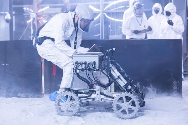 A person in a white hazmat suit and helmet examines a robotic rover on a white, sandy surface at Florida Space Institute Exolith Lab while other figures in hazmat gear observe in the background.