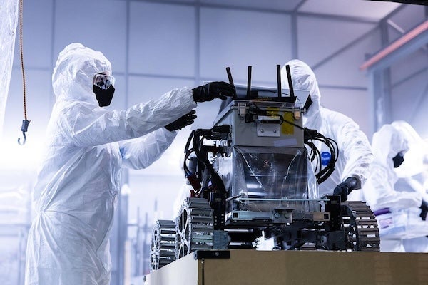 Three people in protective cleanroom suits and masks assemble a robotic rover on a platform during NASA's Lunabotics qualifier at UCF.