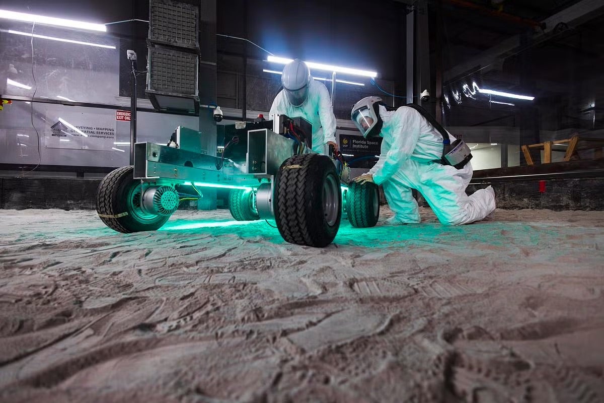 Two people in white protective suits inspect a lunar rover prototype on a simulated moon surface at the Exolith Lab at UCF's Florida Space Institute