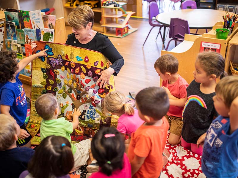 Teacher showing a colorful fabric activity board to children in a classroom.