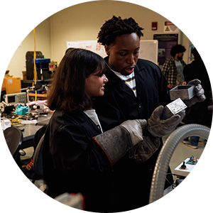 Two students inspect a metal component while wearing protective gear.