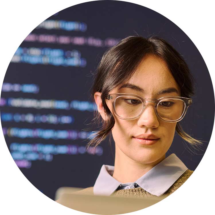Woman wearing glasses works on a computer with programming code in the background.