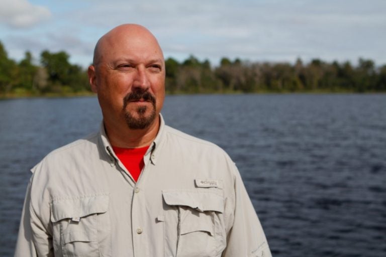 bald man with mustache and khaki button down with red shirt underneath standing by the water with trees in the background