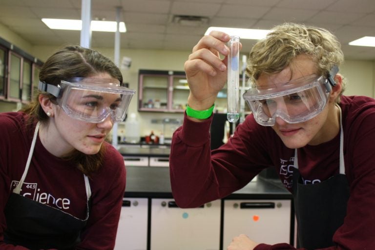 two students in lab with goggles on and maroon long sleeve shirts, looking closely at something