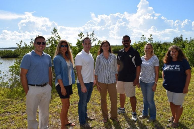 group of 6 people standing side by side smiling at the camera, while they are outside