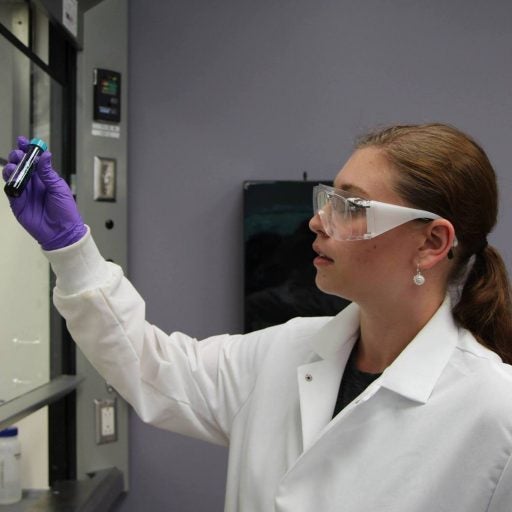 Women in Chemistry - woman with hair pulled back and goggles holding up test tube with purple latex gloves, wearing white lab coat in lab