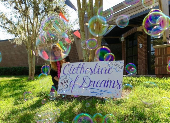 girl sitting outside beside a tree with a sign propped up that says (in cursive) "clothesline of dreams" and there are bubbles flying everywhere
