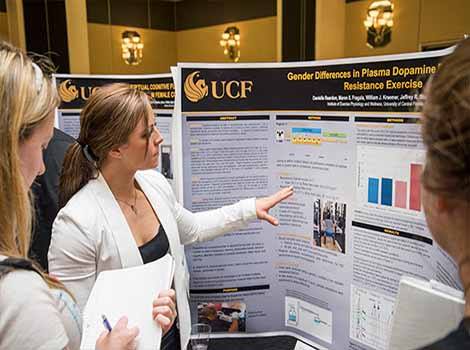 ucf research week, female in white lab coat pointing to trifold posterboard, explaining study to observers