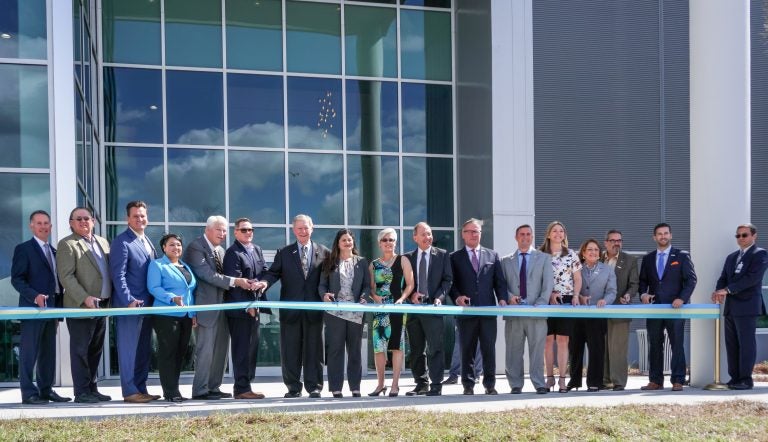 bridge ribbon cutting ceremony. group of men and women in suits standing in front of glass building, about to cut a long ribbon