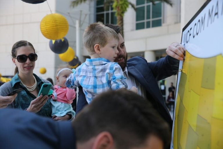 man in suit holding little boy in plaid shirt, theyre taking a selfie in a large crowd with black and gold balloons in the background