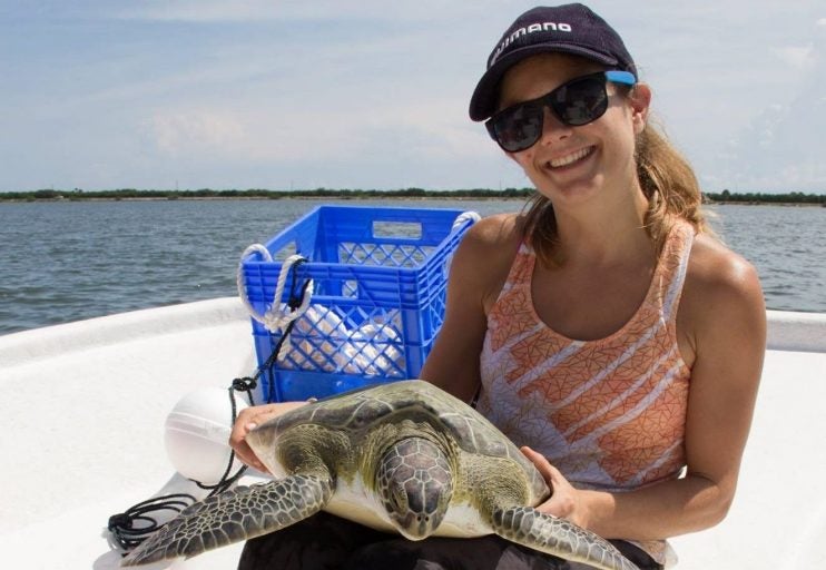 woman with hair pulled back, wearing sunglasses and hat, smiling and holding sea turtle on a boat