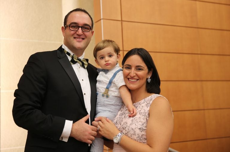 husband in tuxedo and wife in dress holding young boy in polo and suspenders