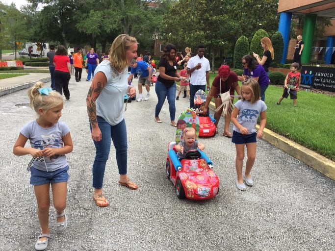 group of adults and children walking on sidewalk, one child in small child-sized red racecar