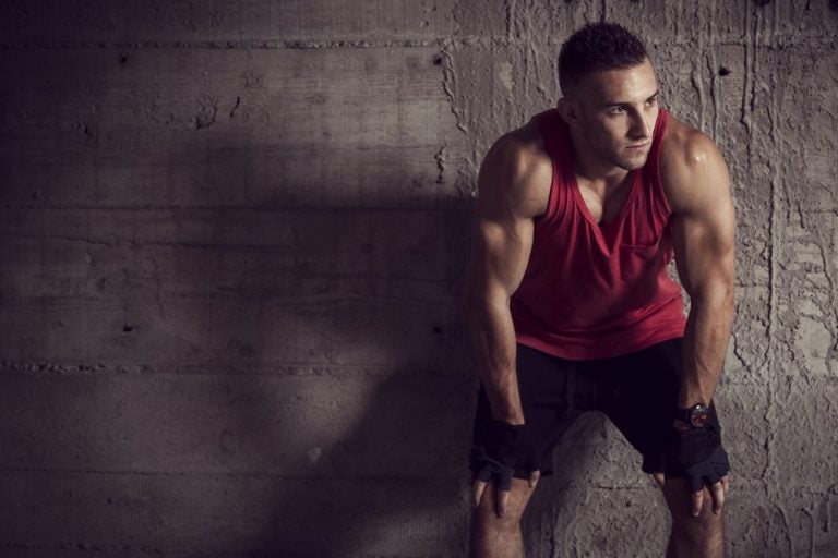 muscular man leaning against wall in black shorts and red tanktop