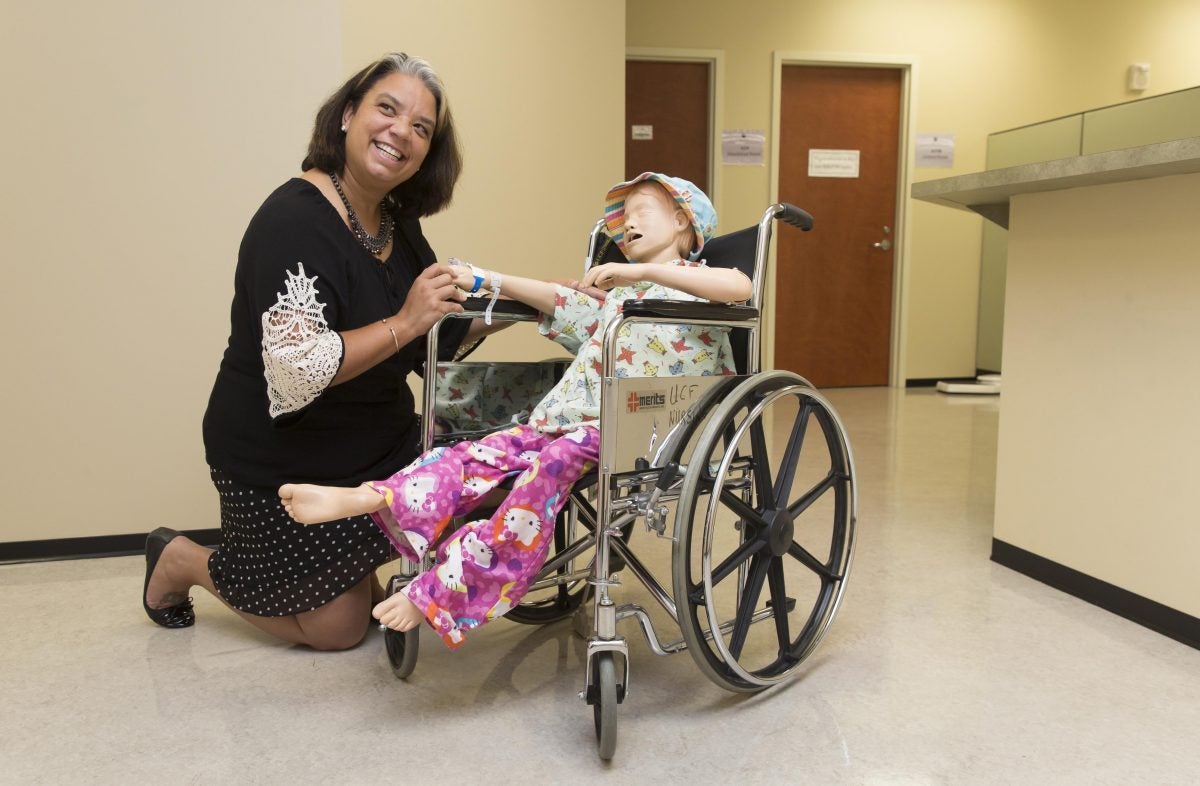 woman in a black dress with white sleeves kneels next to a mannequin in hospital scrubs sitting in a wheelchair