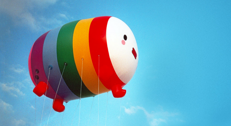 giant balloon, rainbow colored with happy face on the front, flying in blue sky