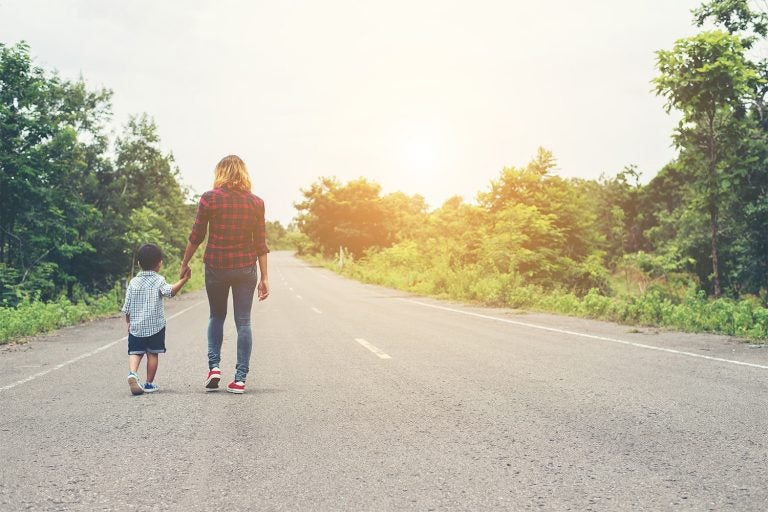 mother and son walking away from camera down a paved road with shubbery on either side