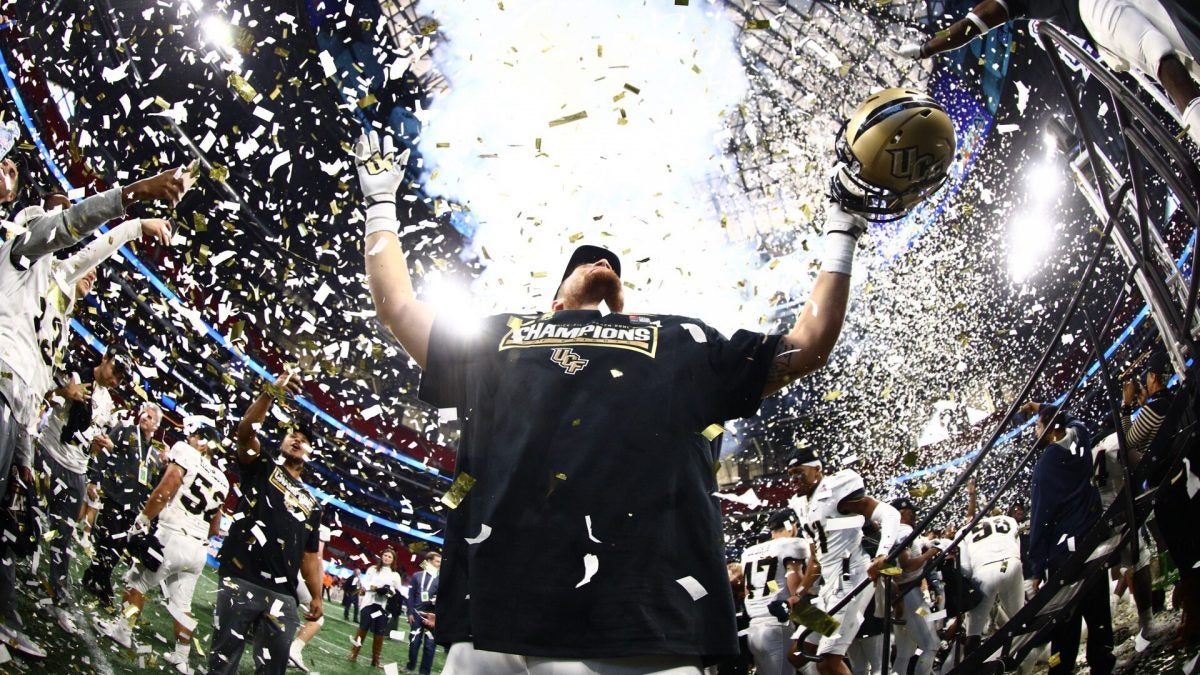 White and gold confetti showers a player wearing a black "Champions" shirt as he holds his gold helmet and looks out at the crowd.