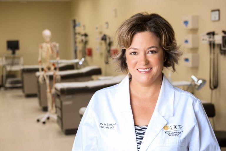 femal ucf doctor with short hair smiling in white lab coat, standing in front of patient beds with fake skeleton in background