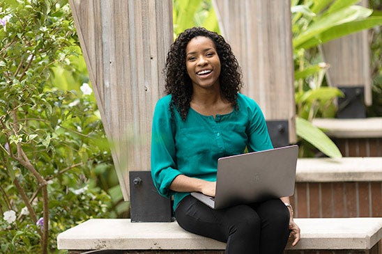 A smiling woman wearing a green top sits outside with a laptop.