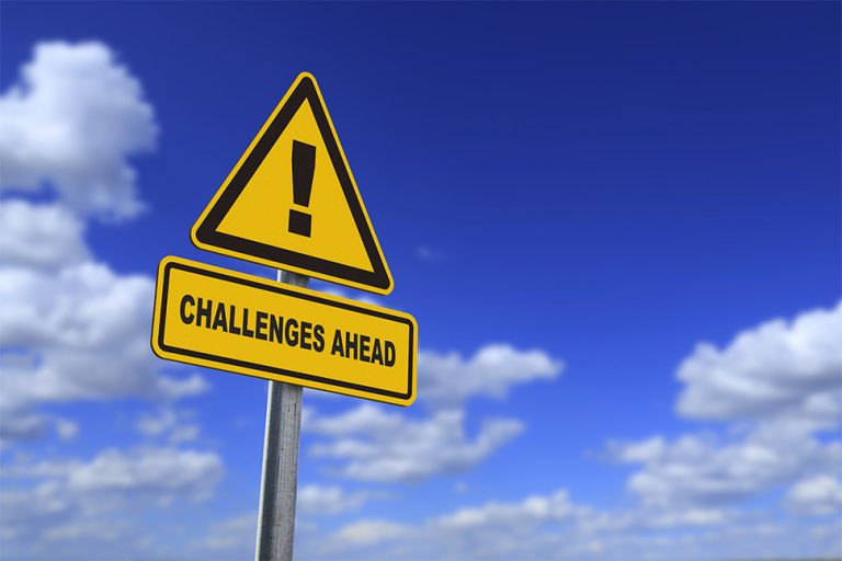 yellow triangular street sign that has an exclamation mark and 'challenges ahead' written in black. in front of a blue sky background with fluffy white clouds.