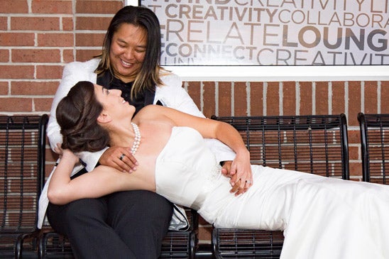 A woman in a wedding gown reclines in the lap of a woman in a black suit with a white jacket.