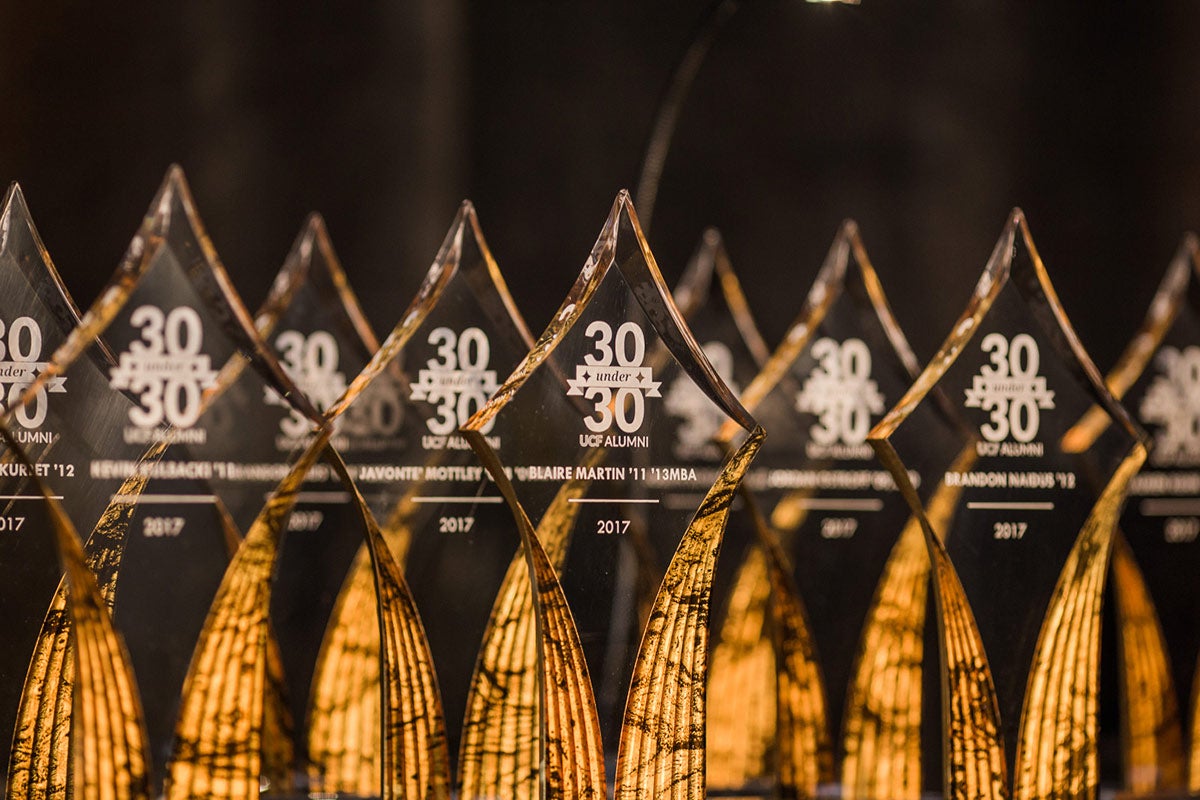 Nine glass awards in the shape of diamonds are in front of a black background and are backlit with yellowish orange color. The awards say 30 Under 30 in white text.