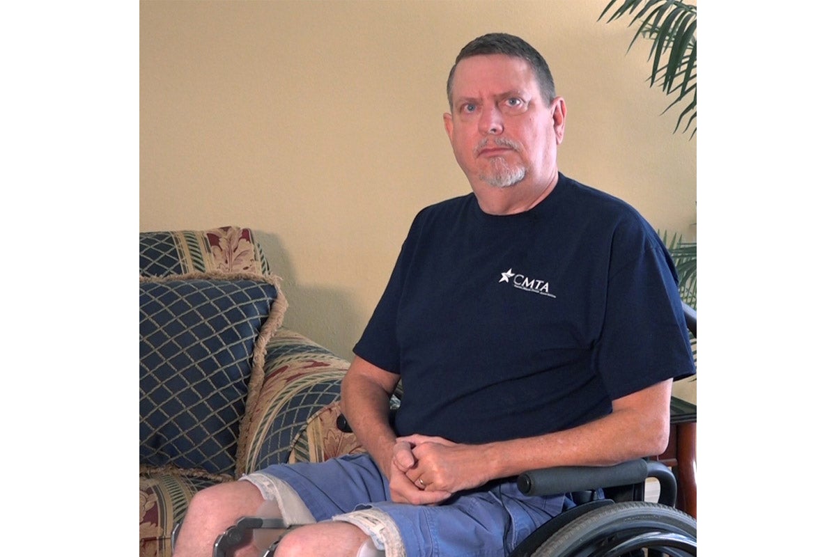 A white man in denim shorts and a navy blue t-shirt sits in a wheelchair, facing the camera.