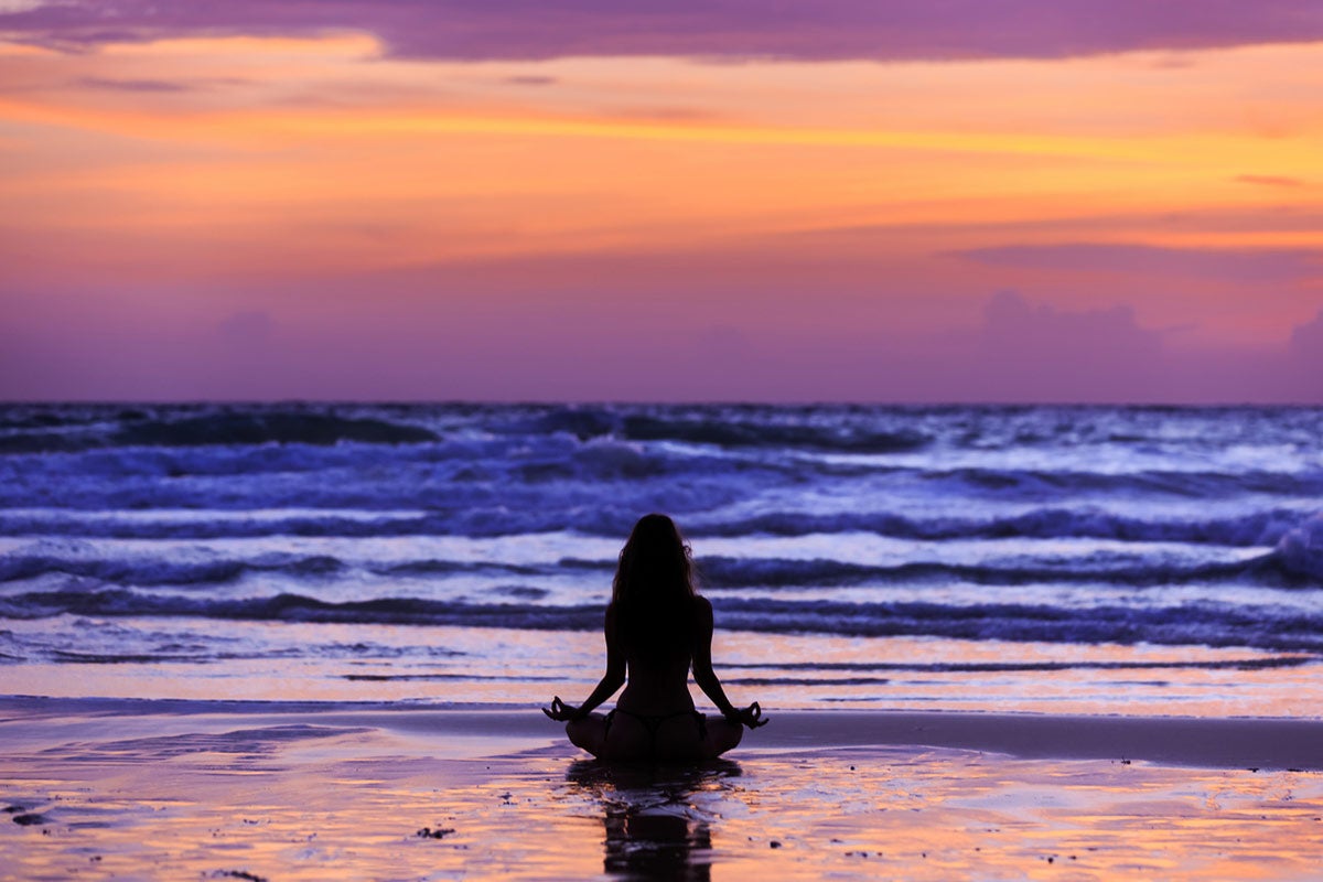 Silouette of a woman seated in lotus position on the beach at sunrise.