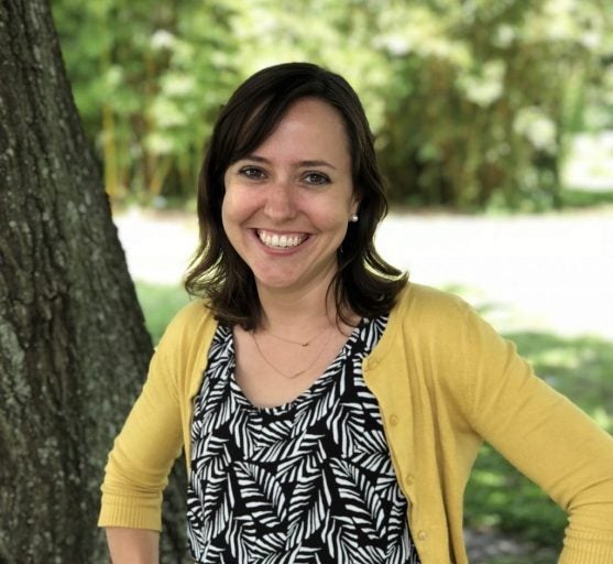 female with brown hair, brown eyes, big smile, yellow sweater over black and white blouse, standing in front of a tree