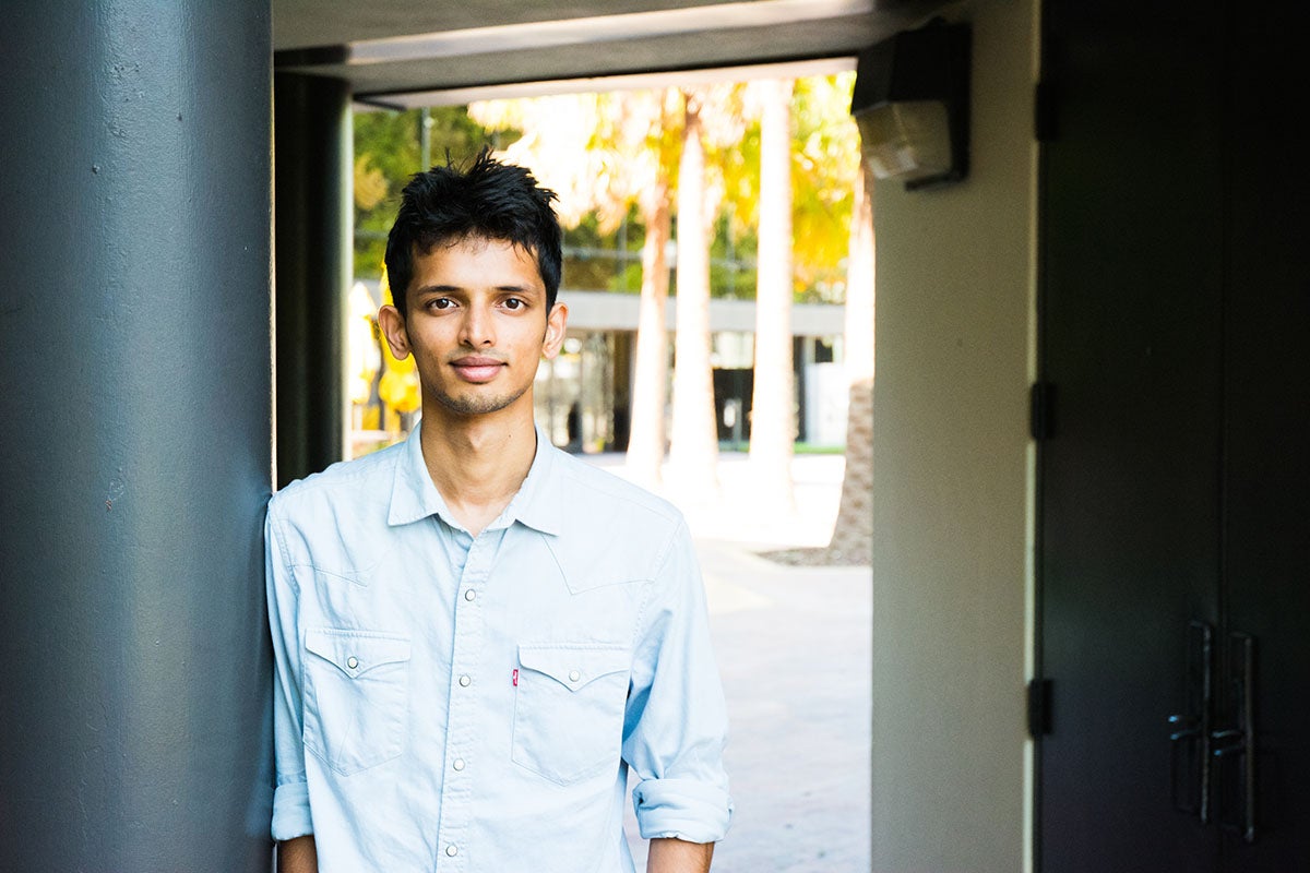 Dark haired Indian student in blue shirt leaning against a building column.