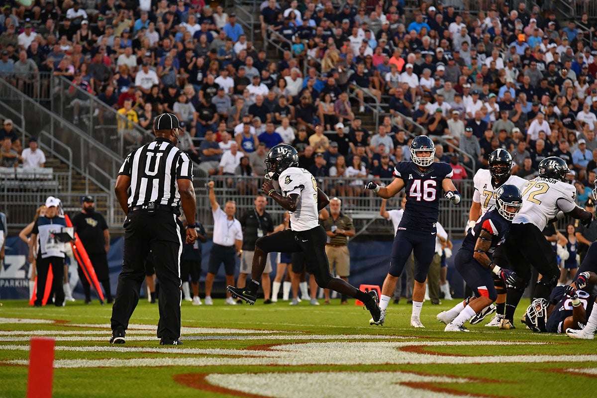 Football player in white jersey and black pants runs into end zone with referee looking on