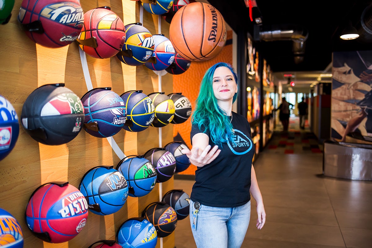 young woman with green and blue long hair, wearing a black t shirt and blue jeans, stands in front of a wall of different color mounted basketballs