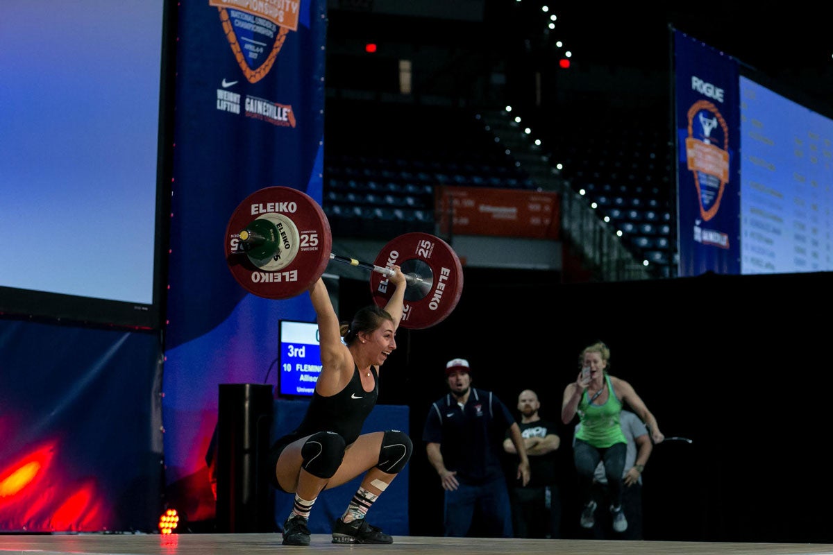 Young adult woman lifts barbell with weights overhead in a squatting position on stage