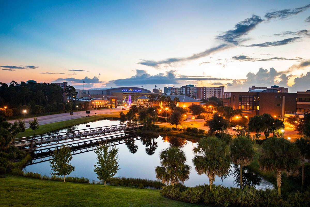 Wide view of campus with a pond in the foreground, amber lights and CFE Arena in the back as the sun rises