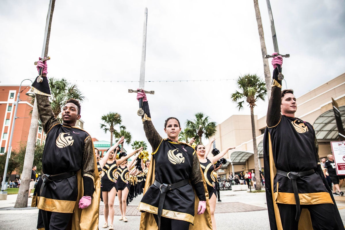 Two men and a woman dressed in black and gold Knight costumes each hold a sword to the sky as they walk down the street with cheerleaders marching behind them.
