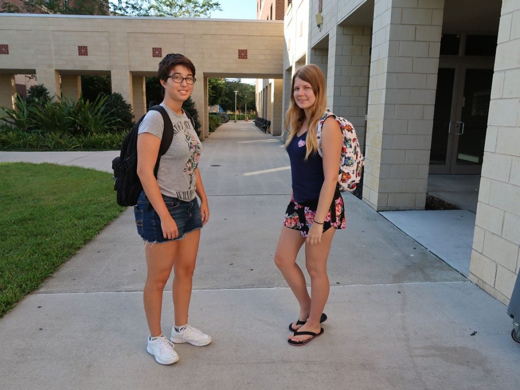 Two female students wearing t shirts and shorts pose on a sidewalk with backpacks over their shoulders