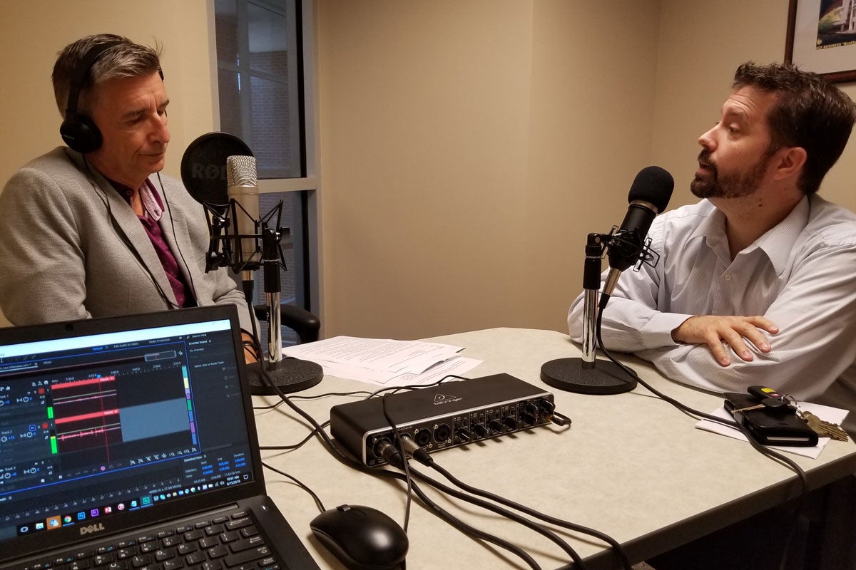 two men in a recording room sit at a table with standup microphones facing each other having a conversation as a laptop captures the audio